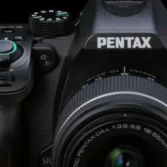 Zbliżenie na czarną lustrzankę Pentax KM z obiektywem 18-55mm. Widoczne pokrętła, przyciski i logo producenta.