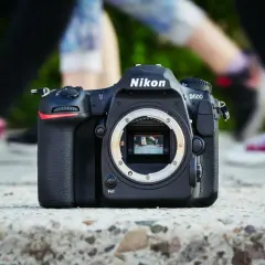Nikon D500 na kamieniu, z rozmazanymi nogami w tle. Dowiedz się, jak sprawdzić przebieg aparatu Nikon.
