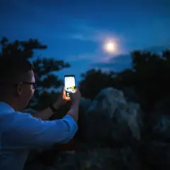 Chłopak robi zdjęcie księżyca telefonem Xiaomi. Nocne niebo, drzewa i skały w tle.