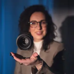 Uśmiechnięta kobieta w okularach unosi obiektyw aparatu nad dłonią. Czy fotograf przechowuje zdjęcia w obiektywie?