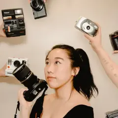 Kobieta z aparatem w ręku, otoczona przez inne aparaty: Polaroid, Nikon, cyfrówki. Idealna do wyboru, jaka lustrzanka na początek.