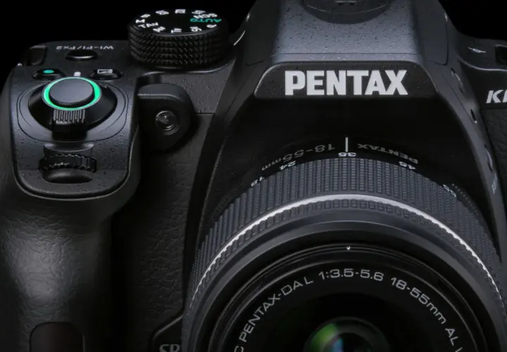Zbliżenie na czarną lustrzankę Pentax KM z obiektywem 18-55mm. Widoczne pokrętła, przyciski i logo producenta.
