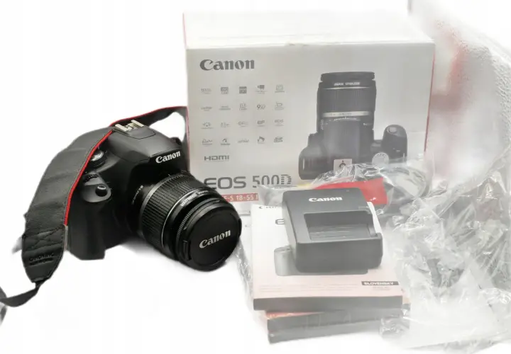 Aparat Canon EOS 500D z obiektywem i akcesoriami na białym tle.