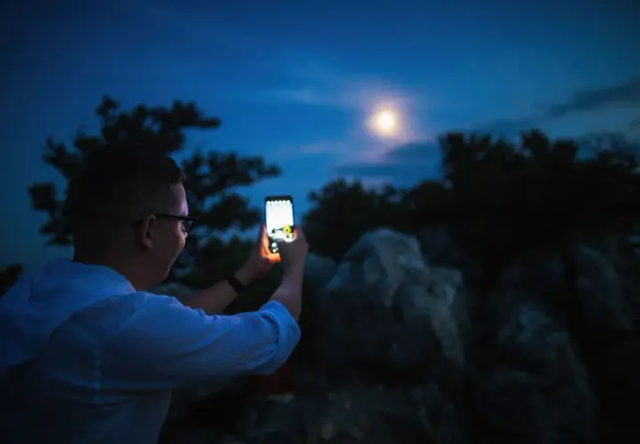 Chłopak robi zdjęcie księżyca telefonem Xiaomi. Nocne niebo, drzewa i skały w tle.
