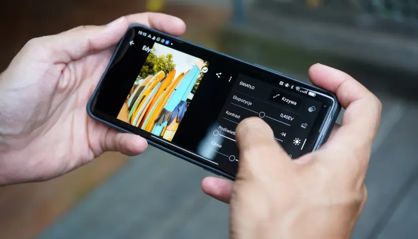 Ręce trzymają smartfon, na którym widać edycję zdjęcia desek surfingowych. Dowiedz się, jak zrobić zdjęcie z filmu na telefonie, edytując je.