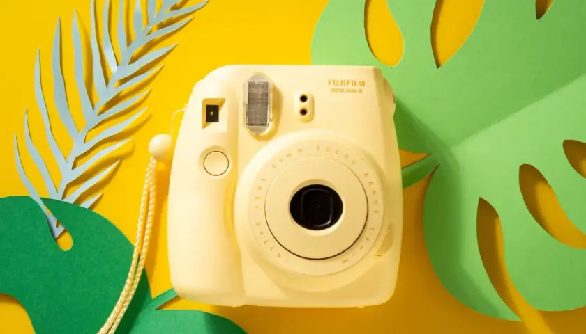 Żółty aparat natychmiastowy Fujifilm Instax Mini 8 na tle tropikalnych liści. Idealny wybór, jeśli zastanawiasz się, jaki aparat natychmiastowy wybrać.