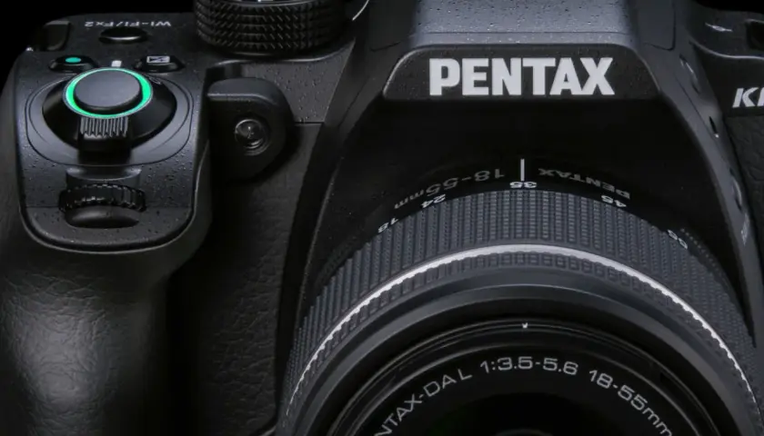 Zbliżenie na czarną lustrzankę Pentax KM z obiektywem 18-55mm. Widoczne pokrętła, przyciski i logo producenta.