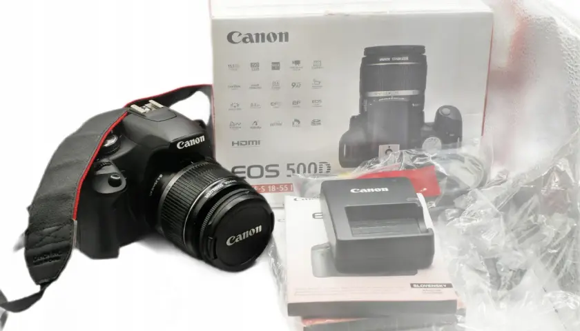 Aparat Canon EOS 500D z obiektywem i akcesoriami na białym tle.