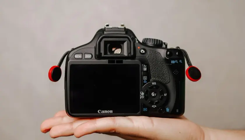 Dłoń trzyma czarną lustrzankę Canon z czerwonymi elementami smyczy.