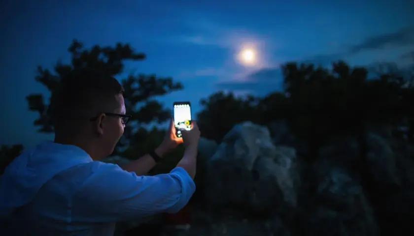 Chłopak robi zdjęcie księżyca telefonem Xiaomi. Nocne niebo, drzewa i skały w tle.