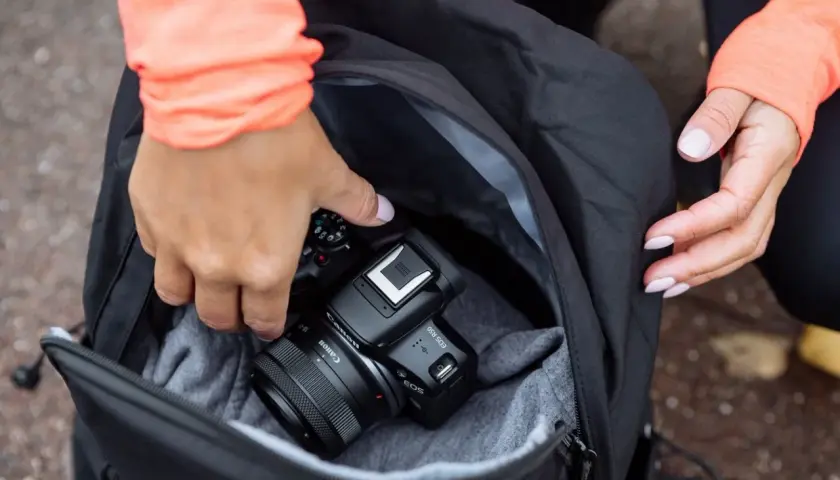 Dłonie wkładają aparat Canon EOS R50 do plecaka. To świetny aparat fotograficzny na początek przygody z fotografią.