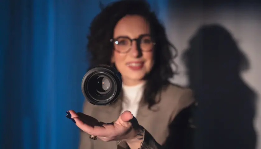 Uśmiechnięta kobieta w okularach unosi obiektyw aparatu nad dłonią. Czy fotograf przechowuje zdjęcia w obiektywie?