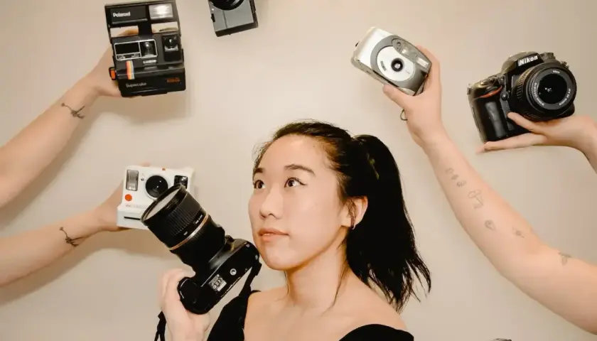 Kobieta z aparatem w ręku, otoczona przez inne aparaty: Polaroid, Nikon, cyfrówki. Idealna do wyboru, jaka lustrzanka na początek.