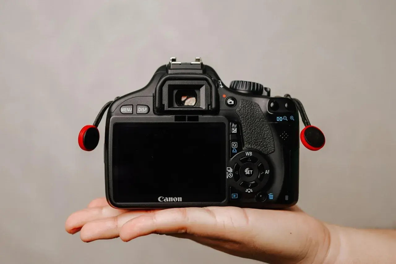 Dłoń trzyma czarną lustrzankę Canon z czerwonymi elementami smyczy.