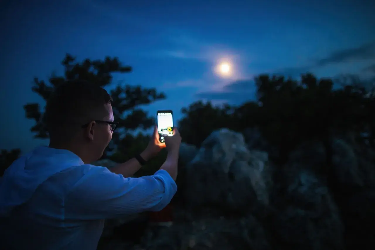 Chłopak robi zdjęcie księżyca telefonem Xiaomi. Nocne niebo, drzewa i skały w tle.