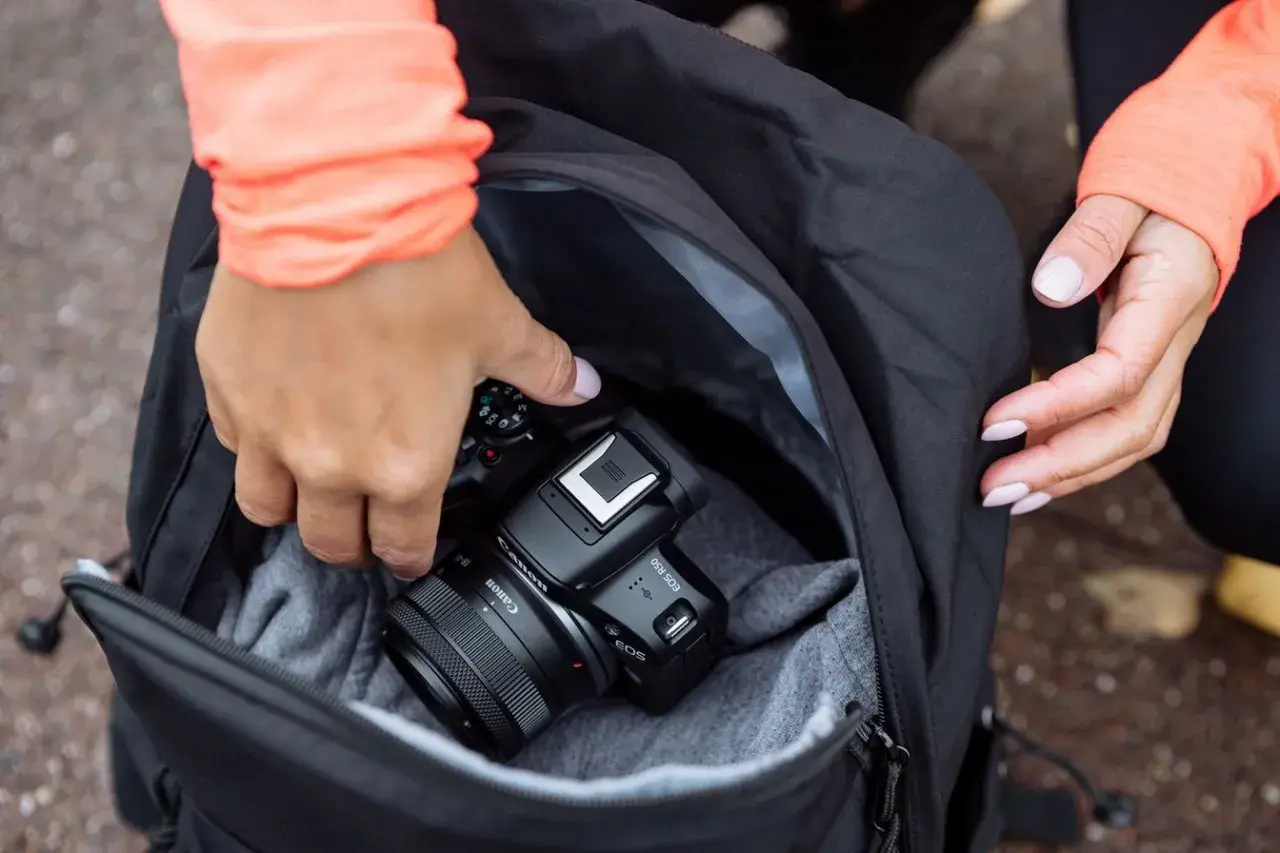 Dłonie wkładają aparat Canon EOS R50 do plecaka. To świetny aparat fotograficzny na początek przygody z fotografią.