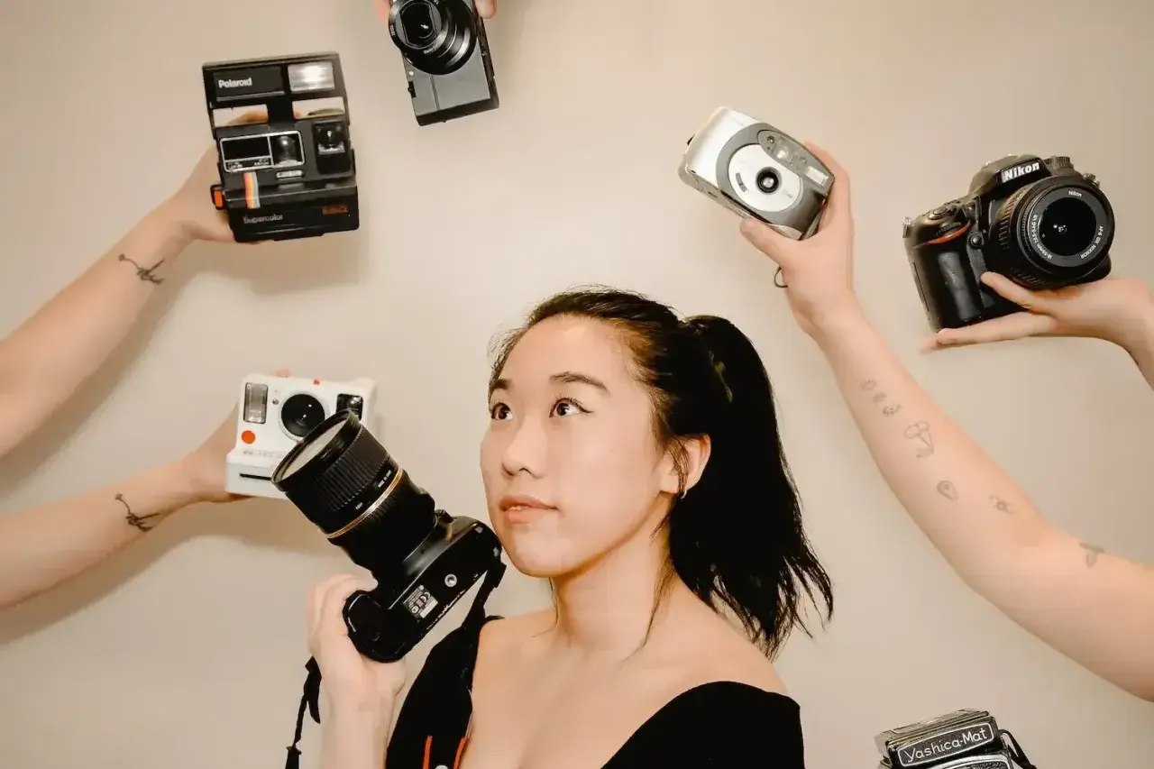 Kobieta z aparatem w ręku, otoczona przez inne aparaty: Polaroid, Nikon, cyfrówki. Idealna do wyboru, jaka lustrzanka na początek.