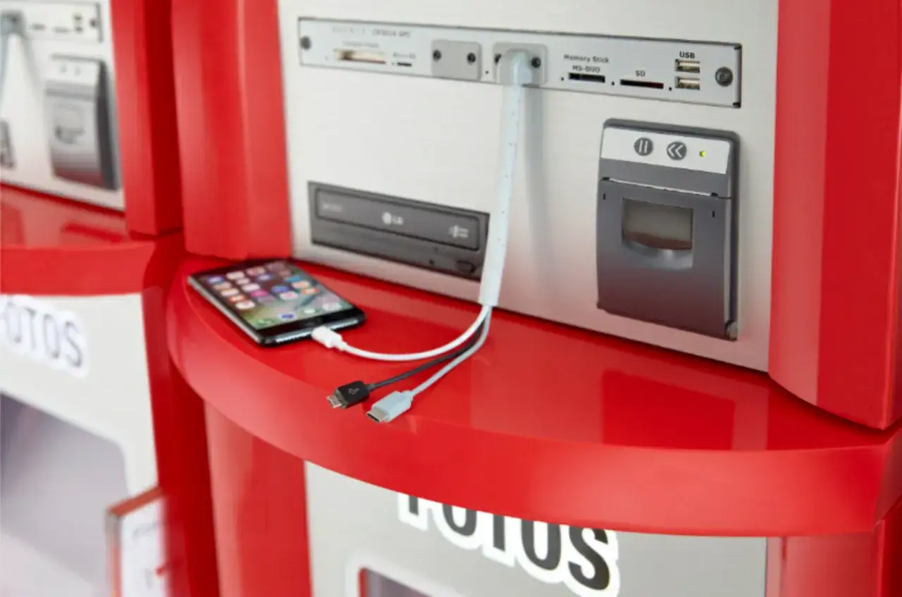 Czy można wywołać zdjęcia z telefonu u fotografa? Tak, ten automat z czerwonymi elementami i ekranem telefonu podłączonym kablem sugeruje taką możliwość.