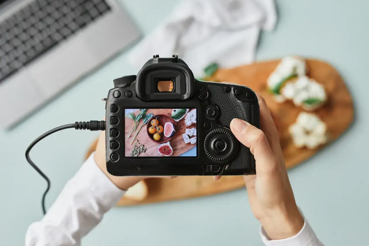 Ręce trzymają aparat fotograficzny lustrzanka, na ekranie widać kompozycję z pomidorami i figami.
