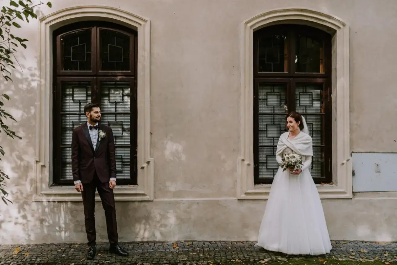 Para o ślubu, zapytaj fotografa o uchwycenie tych chwil. Panna młoda w białej sukni i pan młody w bordowym garniturze pozują przed oknami.