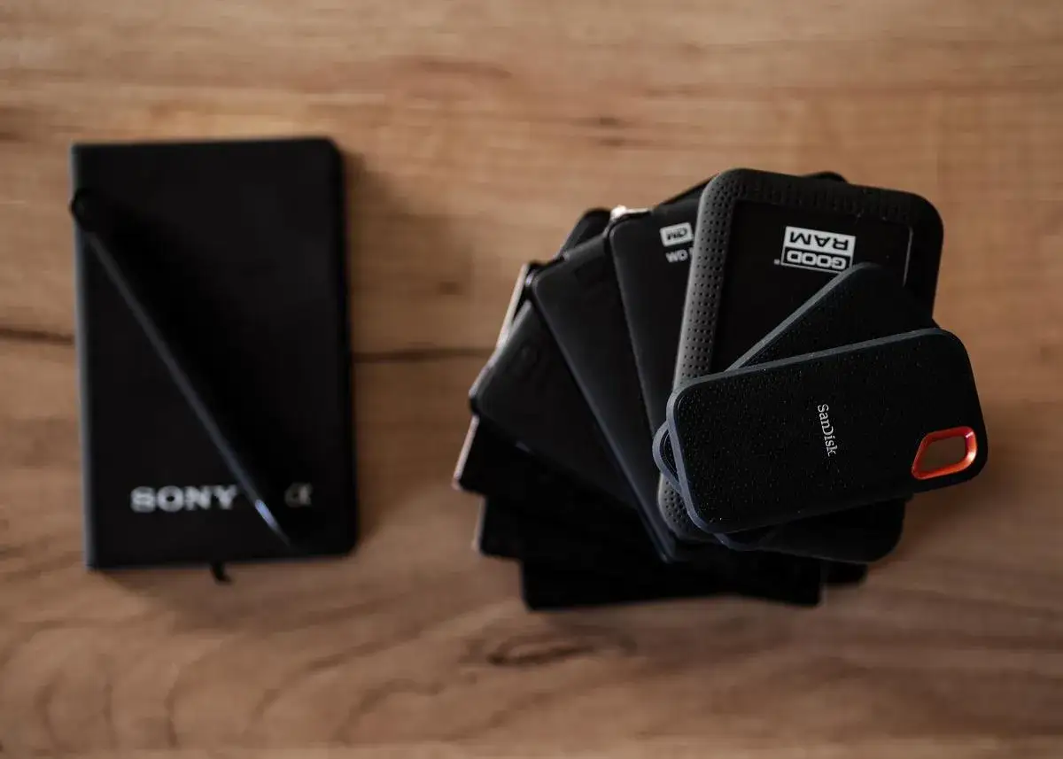 Na drewnianym blacie leży czarny notatnik Sony i stos zewnętrznych dysków twardych, na których fotograf przechowuje zdjęcia.