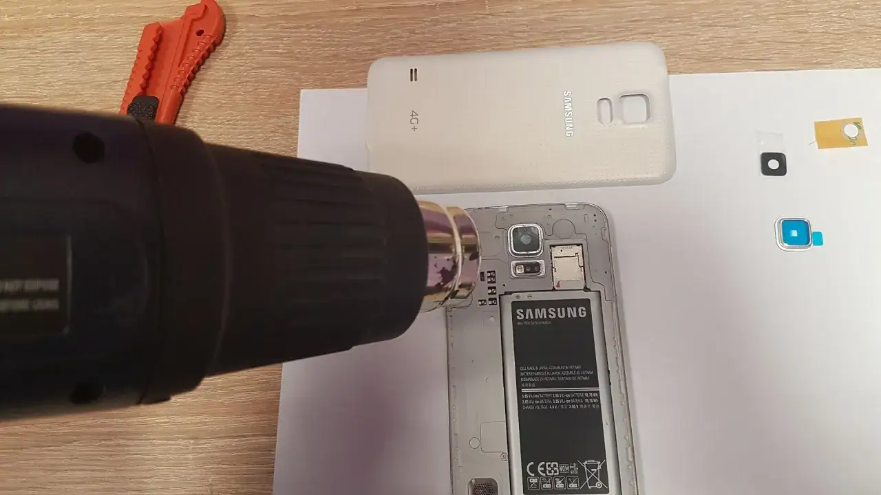 Ciepłe powietrze z opalarki ułatwia demontaż, gdy chcesz wymienić szybkę aparatu w telefonie. Obok leży rozkręcony Samsung.