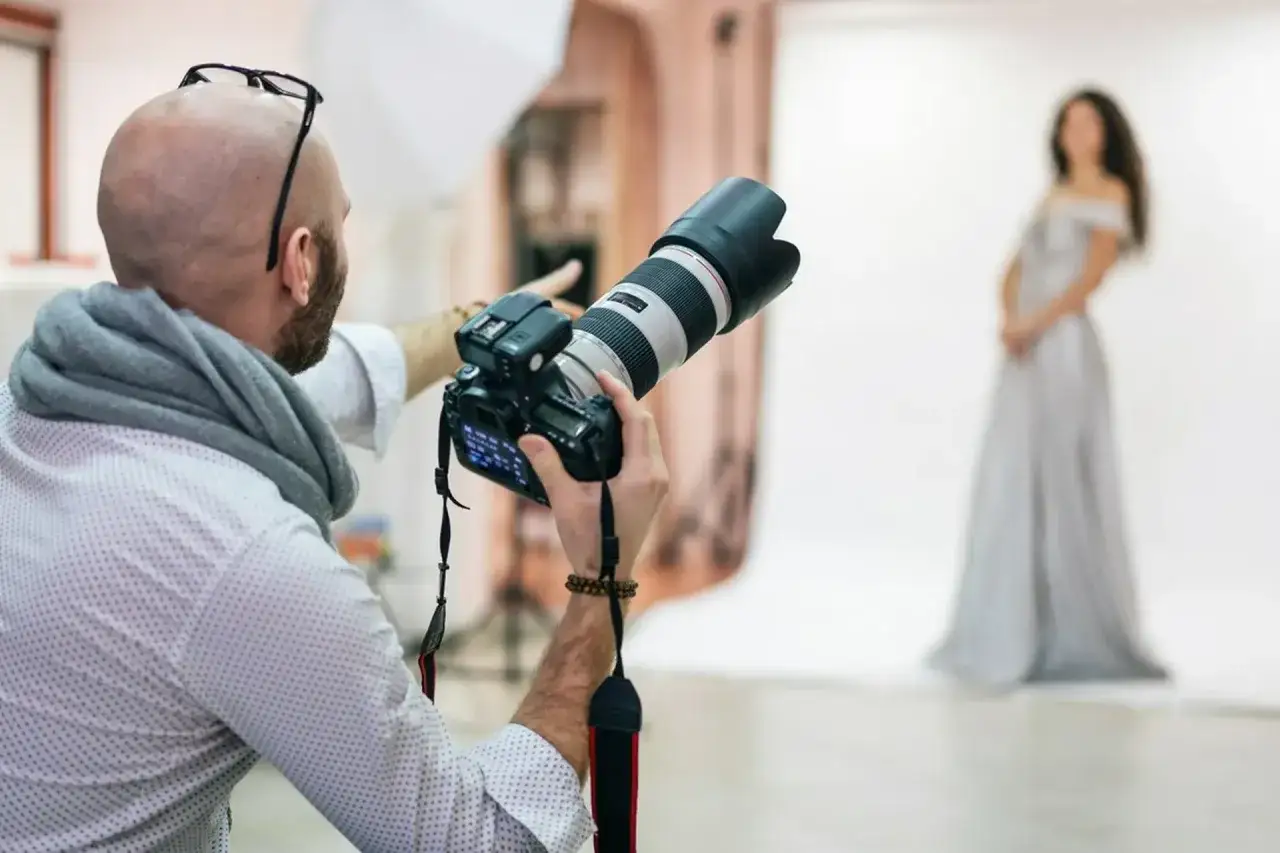 Fotograf w białej koszuli i szarym szalu kieruje obiektyw na modelkę w jasnej sukni. Zastanawiasz się, ile kosztuje sesja u fotografa?